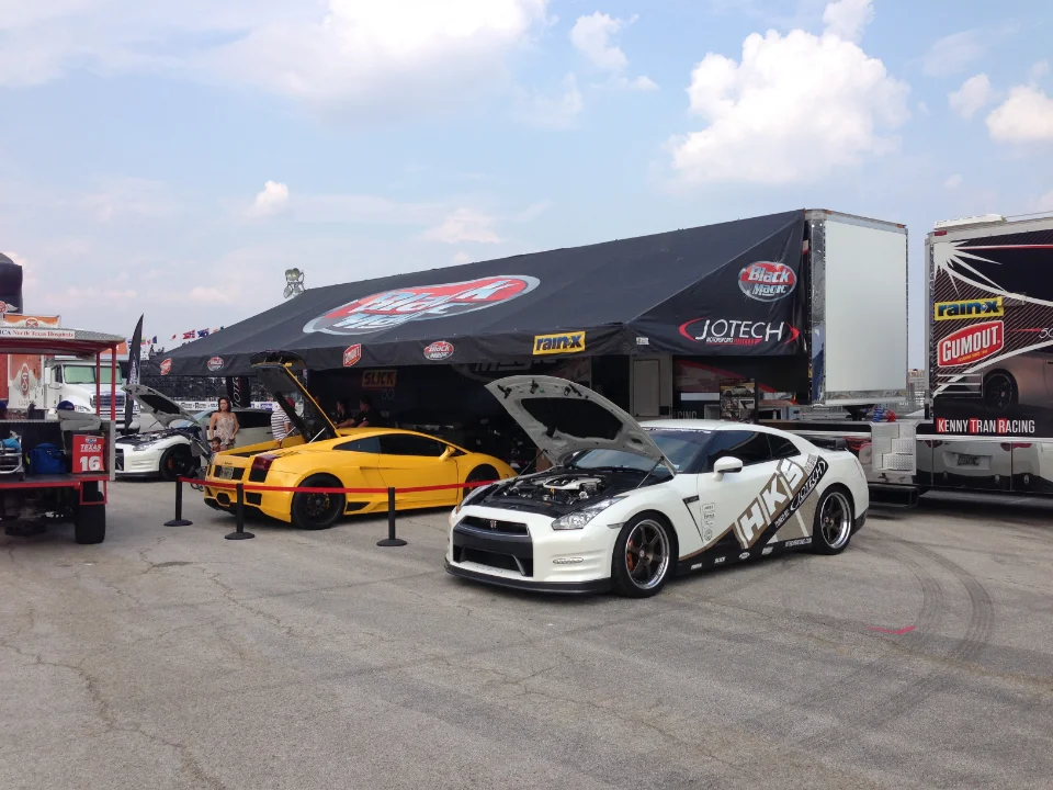 A white Nissan GT-R and a yellow Lamborghini Gallardo with open hoods displayed under a Jotech Motorsports tent at an outdoor car event.
