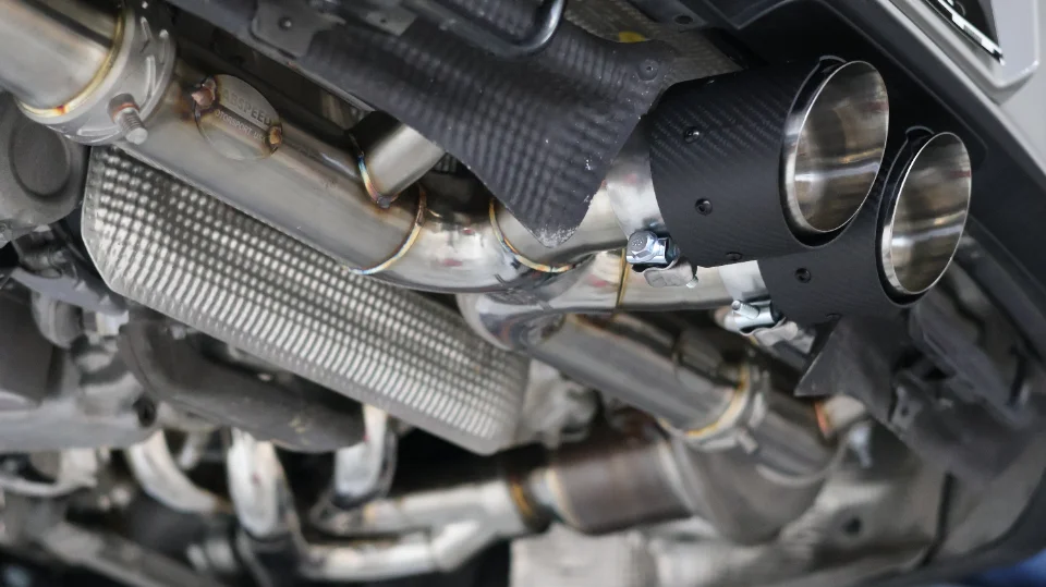 Close-up view of a performance exhaust system with carbon fiber tips and stainless steel piping installed on a car's underbody.