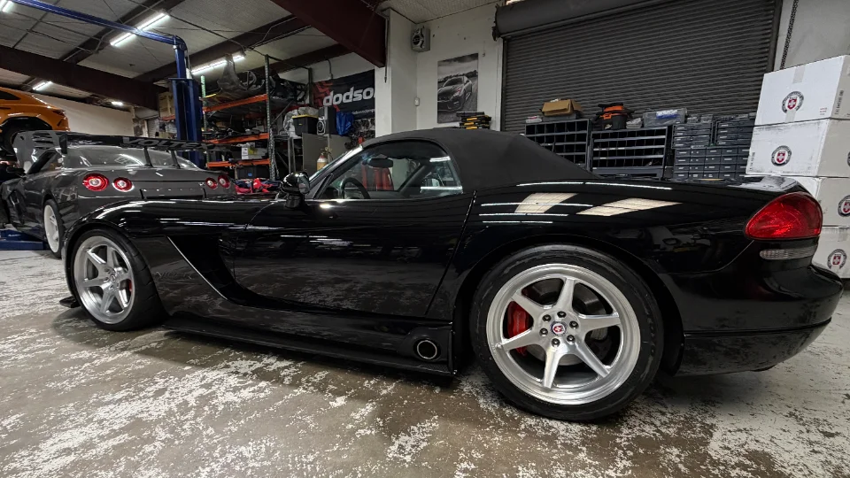 A black Dodge Viper convertible parked on a shop floor with a gray Nissan GT-R on a lift in the background.