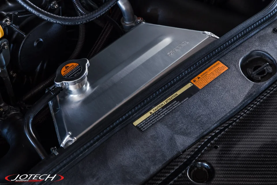 Close-up of an aftermarket aluminum coolant expansion tank with a radiator cap and carbon fiber trim in a car's engine bay.