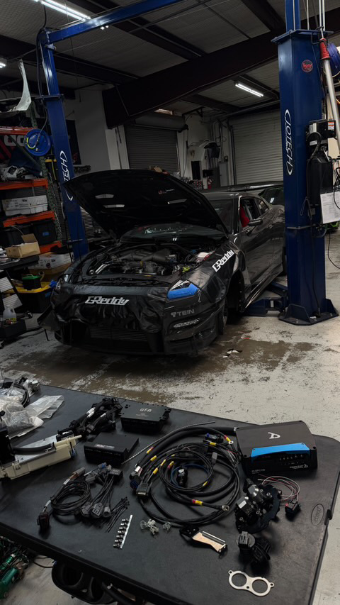 A Nissan GT-R with its hood open and front bumper removed on a lift in a shop, with various wiring harnesses and electronic tuning components laid out on a tabl
