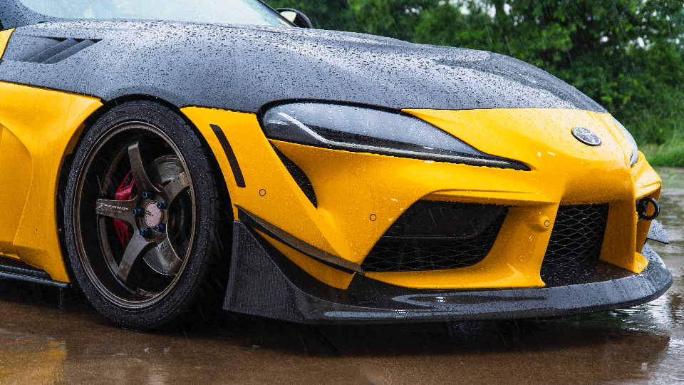 A yellow Toyota Supra with a carbon fiber hood and front splitter, covered in raindrops, showcasing aftermarket wheels.