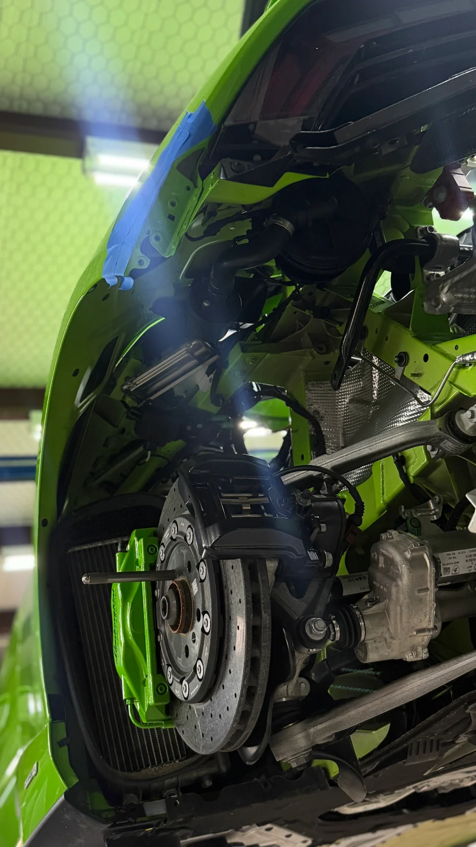 A close-up view of a green Lamborghini's front wheel well, showing the brake caliper, rotor, and suspension components during a shop install.