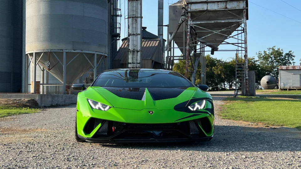 A bright green Lamborghini Huracan with black accents parked on a gravel road in front of large grain silos under a clear sky.