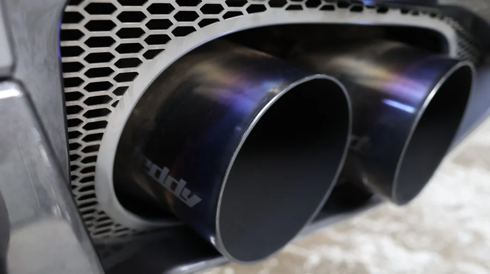 Close-up of Greddy titanium exhaust tips with a honeycomb grille on a performance car's rear bumper.