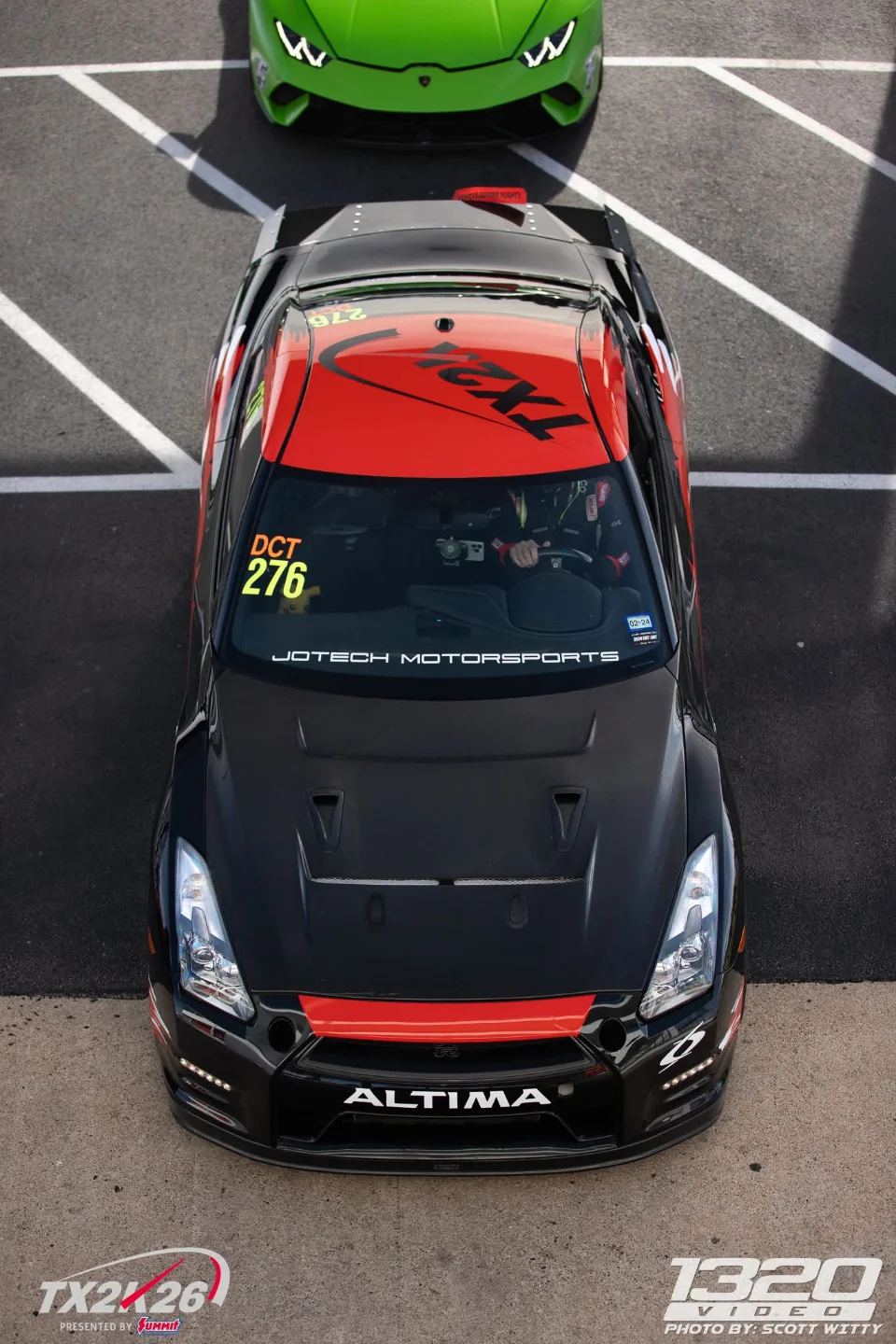A black and red Nissan GT-R with race number 276 in a staging lane at TX2K, with a green Lamborghini in the background.