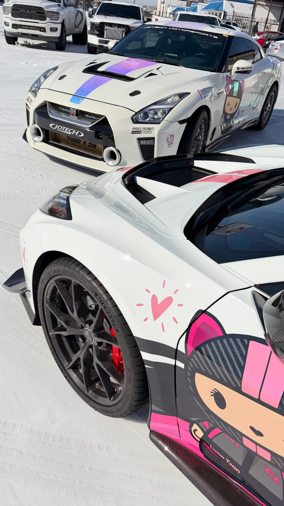 A white Nissan GT-R and a white Corvette with custom pink and purple liveries, featuring Jotech branding, parked in a snowy outdoor setting.