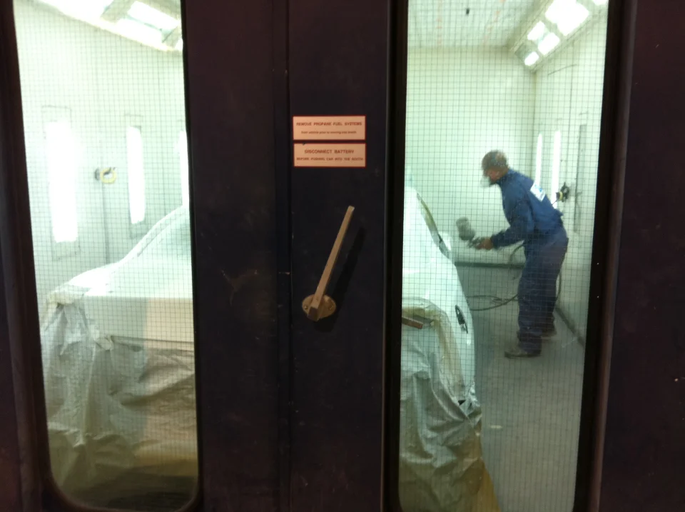 A worker in a blue jumpsuit and respirator sprays a car covered in a tarp inside a paint booth, viewed through a grid-patterned window.