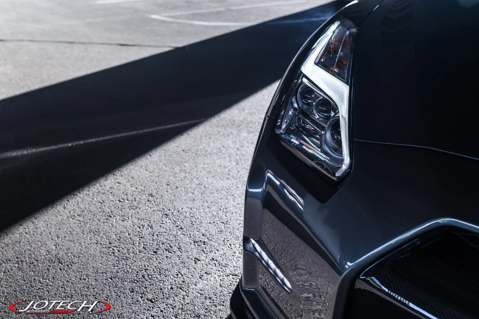 A close-up detail shot of the front headlight and bumper of a dark Nissan GT-R on asphalt with dramatic lighting.
