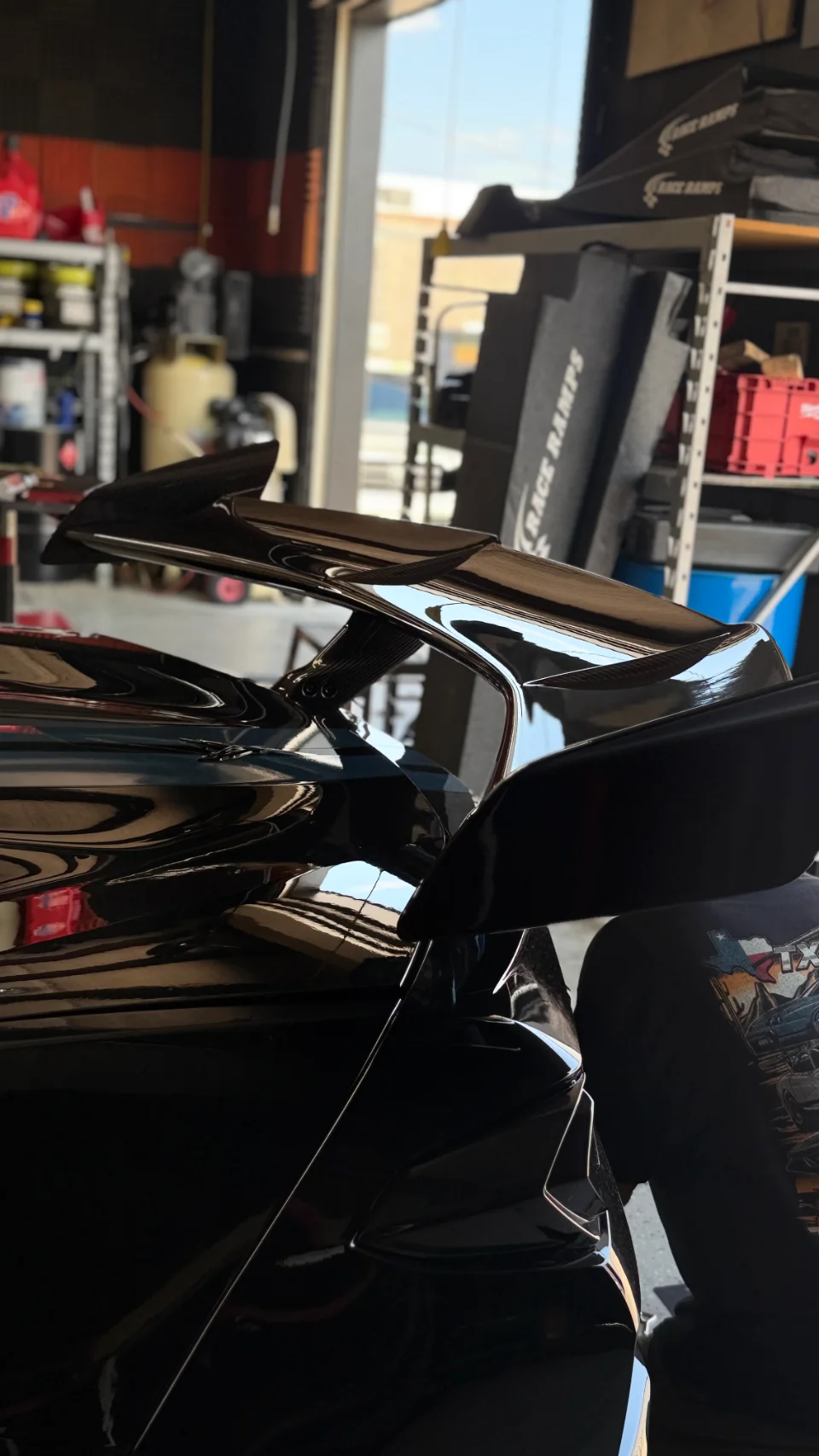 Close-up of a glossy black carbon fiber rear wing on a performance car in a shop setting.