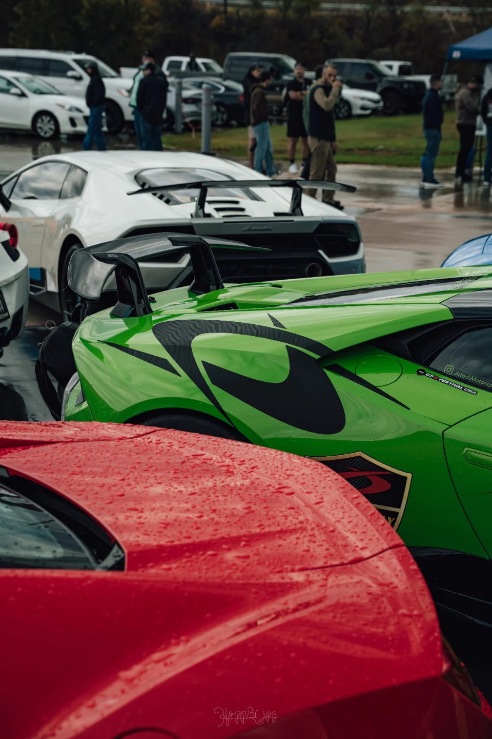 Rear view of a green Lamborghini with a large wing, a white Lamborghini, and a red supercar parked at an outdoor car meet.