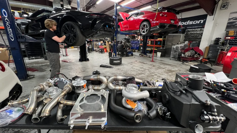 A mechanic works on a black Nissan GT-R on a lift in a performance shop, with a table of turbo and exhaust parts in the foreground.