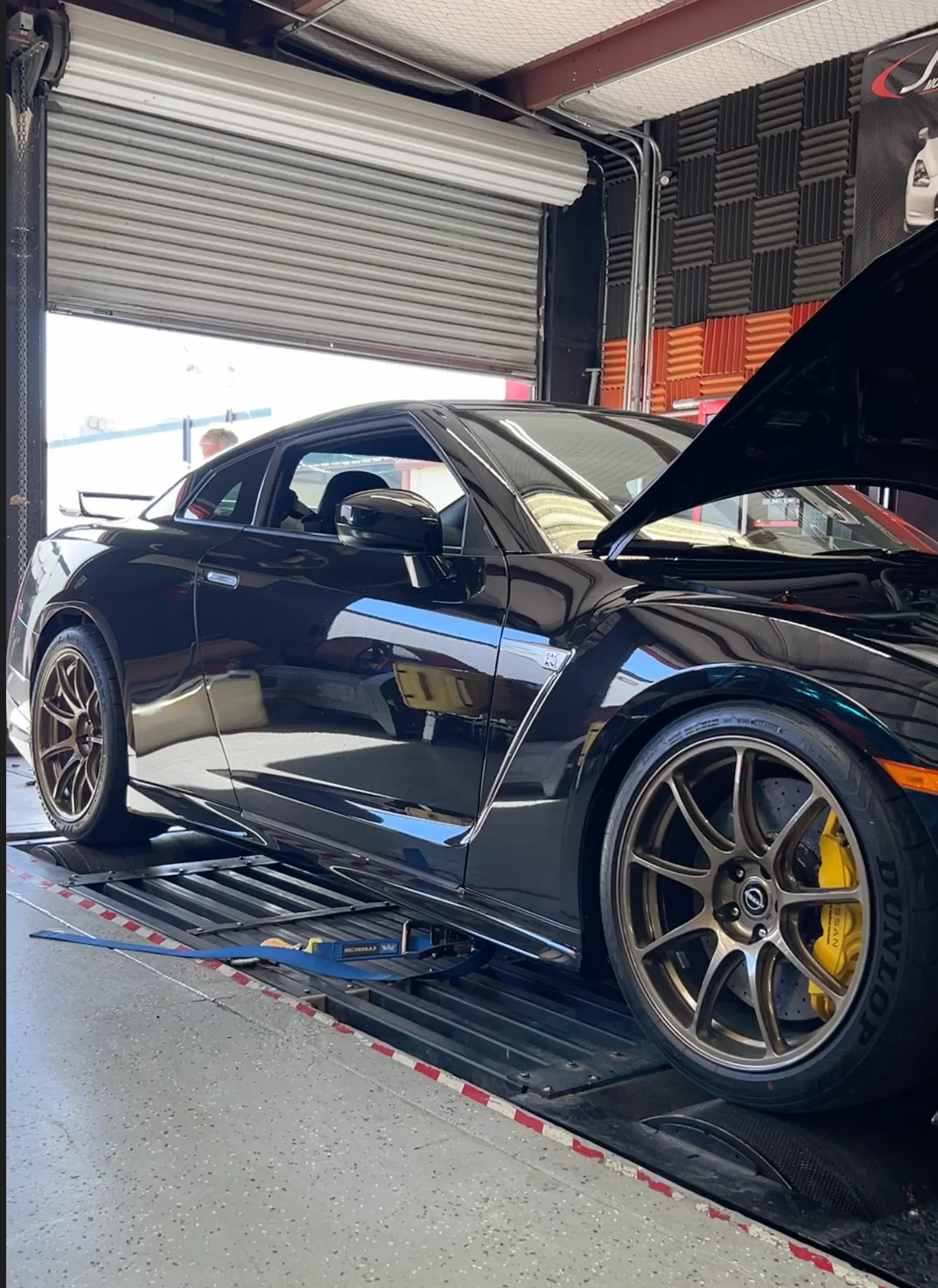A black Nissan GT-R R35 with bronze wheels and yellow calipers strapped to a dyno at a performance shop.