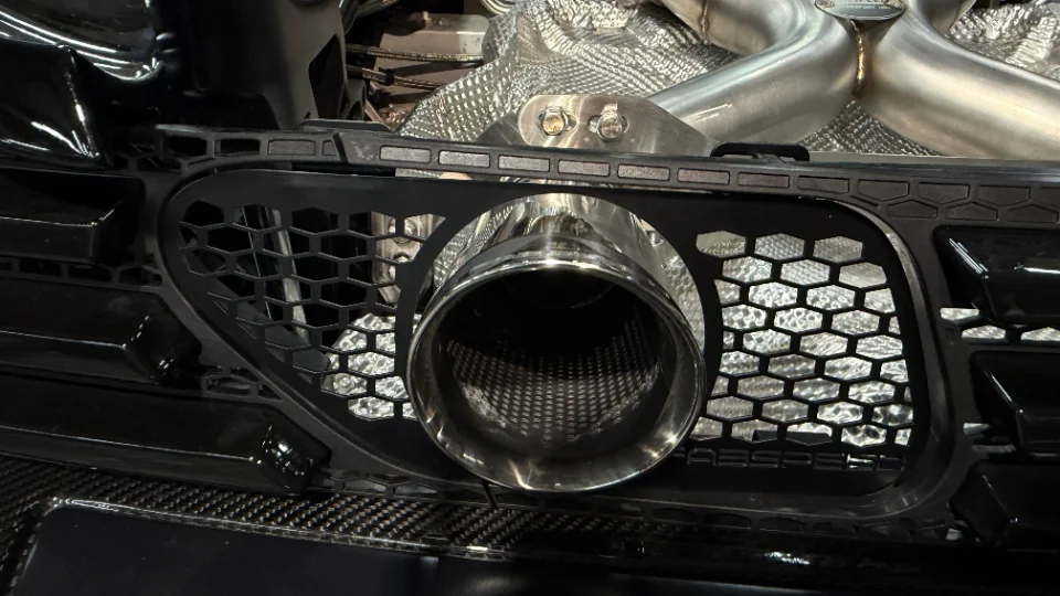Close-up of a polished stainless steel exhaust tip integrated into a black honeycomb grille with carbon fiber trim.