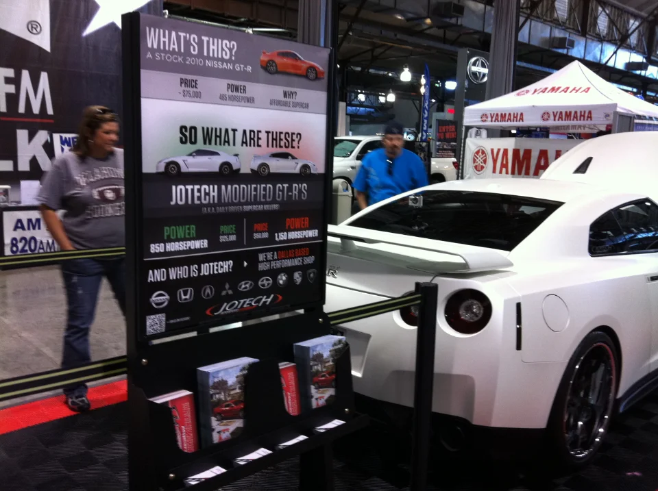A white Nissan GT-R on display at a car show, with a Jotech Motorsports banner promoting modified GT-Rs.