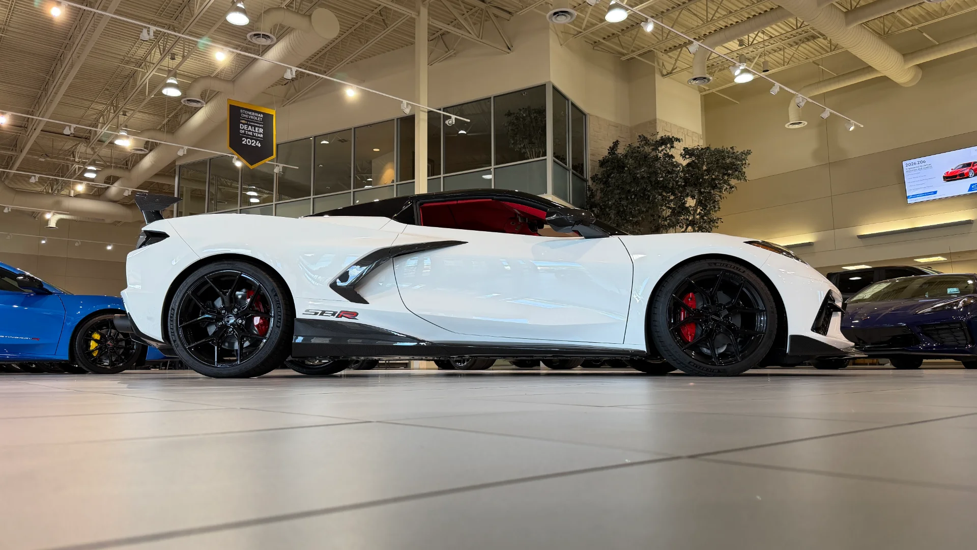 A white Chevrolet Corvette C8 Z06 with a black roof and red interior, parked in a brightly lit car dealership showroom.