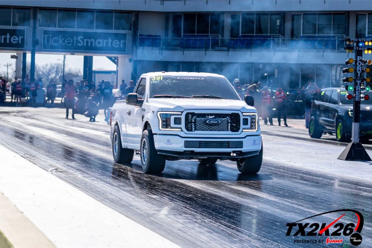 A white Ford F-150 pickup truck performs a burnout at the starting line of a drag strip, with smoke billowing around it.