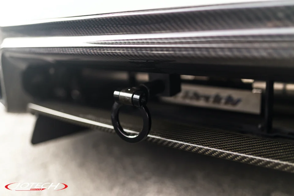 A close-up detail of a carbon fiber rear bumper with a black tow hook on a performance car, featuring Jotech branding.
