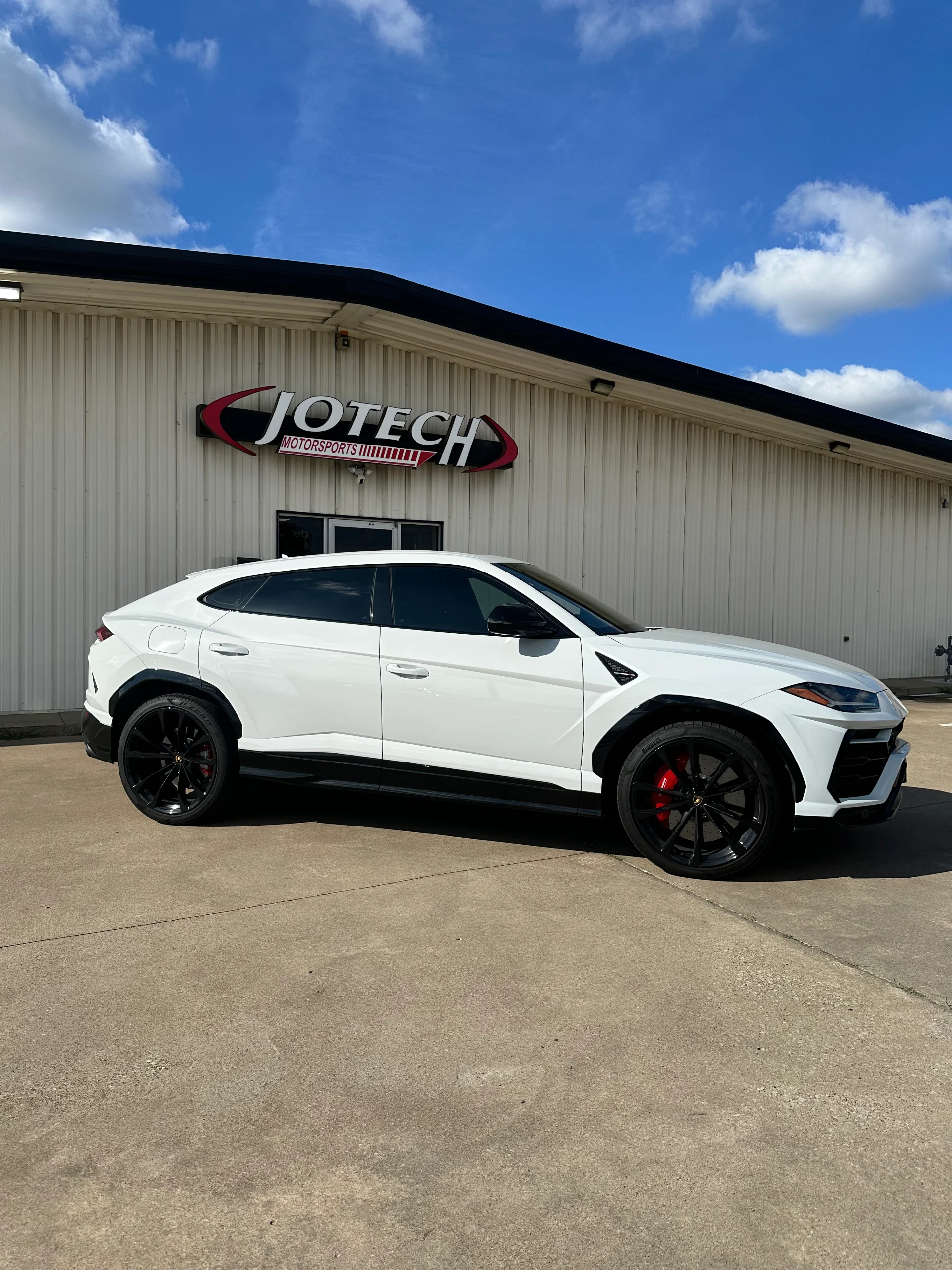 A white Lamborghini Urus luxury SUV with black wheels and red brake calipers parked outside the Jotech Motorsports facility.