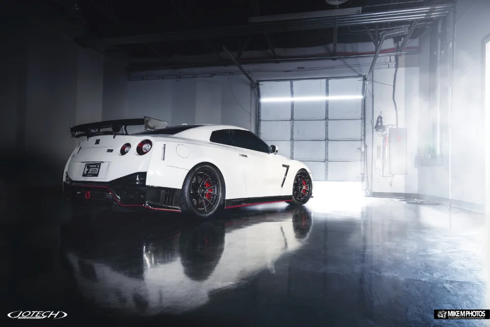 A white Nissan GT-R with aftermarket wheels and a large rear wing parked in a brightly lit garage with a roll-up door.