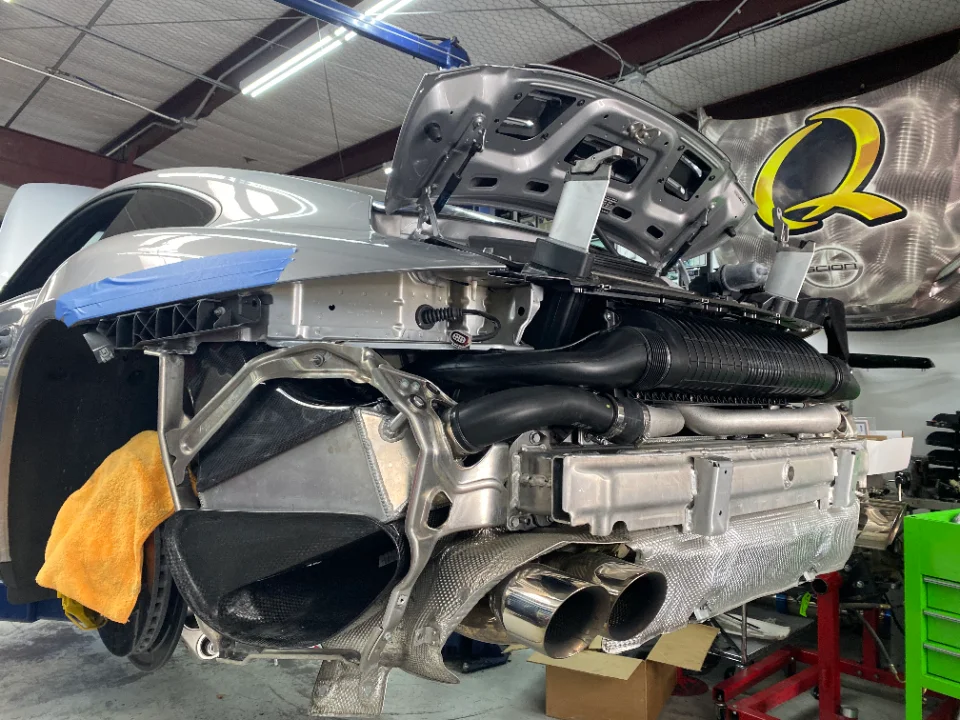 A silver Porsche 911 with its rear bumper removed, exposing the exhaust system, intercoolers, and engine components in a shop.