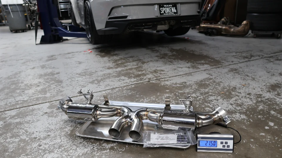 A new stainless steel exhaust system sits on a scale in a performance shop, with a gray Chevrolet Camaro on a lift in the background.