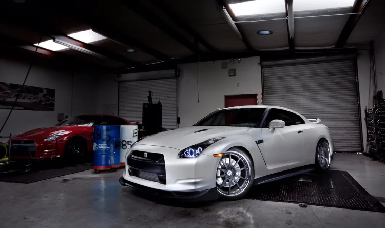 Two Nissan GT-Rs, one white and one red, inside a performance tuning shop with a dyno in the background.