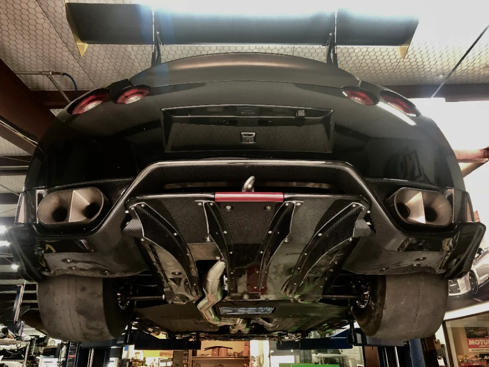Rear view of a black Nissan GT-R on a lift in a performance shop, showcasing a large carbon fiber diffuser, rear wing, and drag tires.