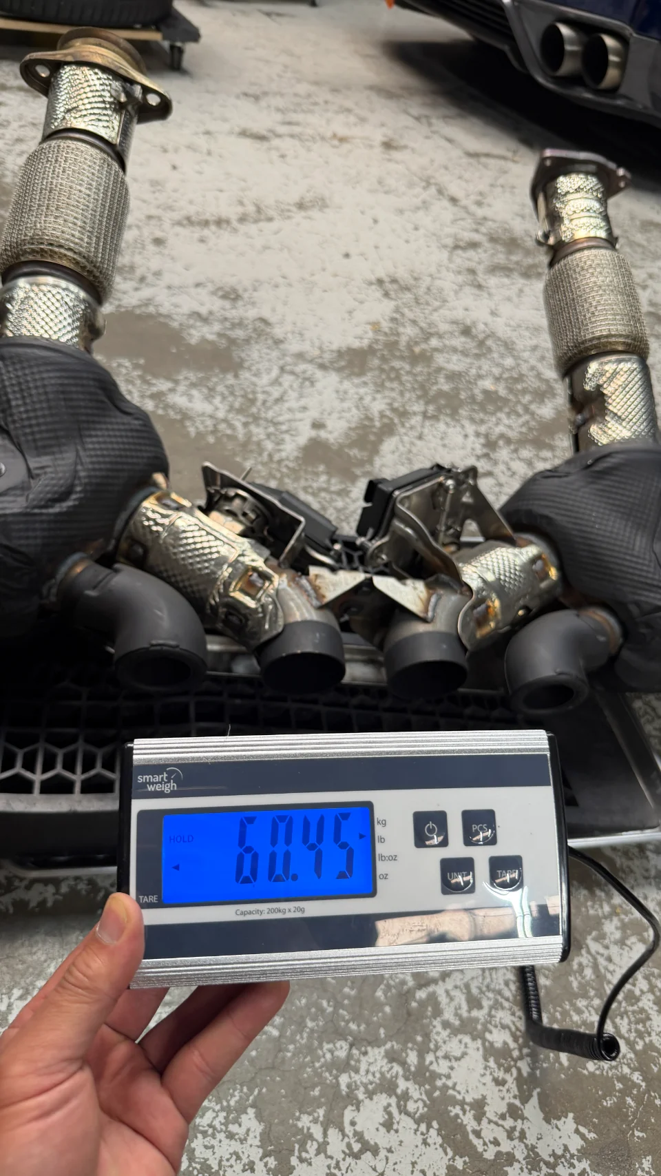 A person weighing performance exhaust downpipes on a digital scale in a shop, showing a reading of 60.45.