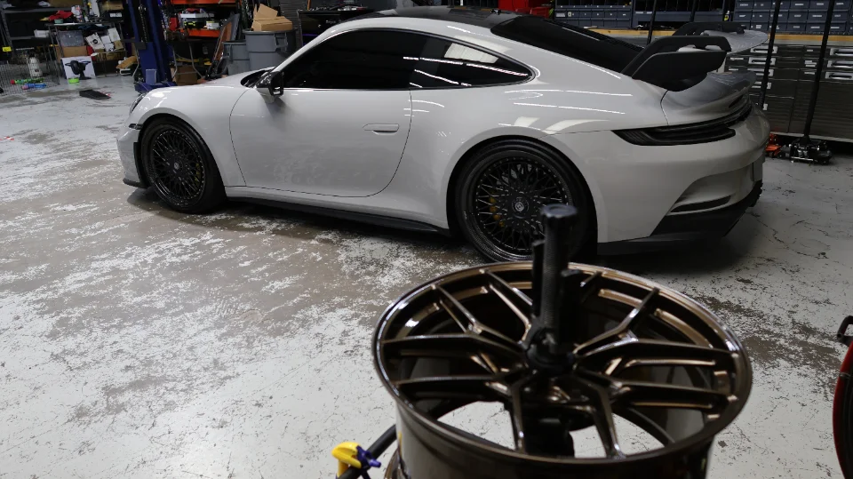 A white Porsche 911 GT3 with aftermarket aftermarket wheels in a performance tuning shop, with a wheel in the foreground.