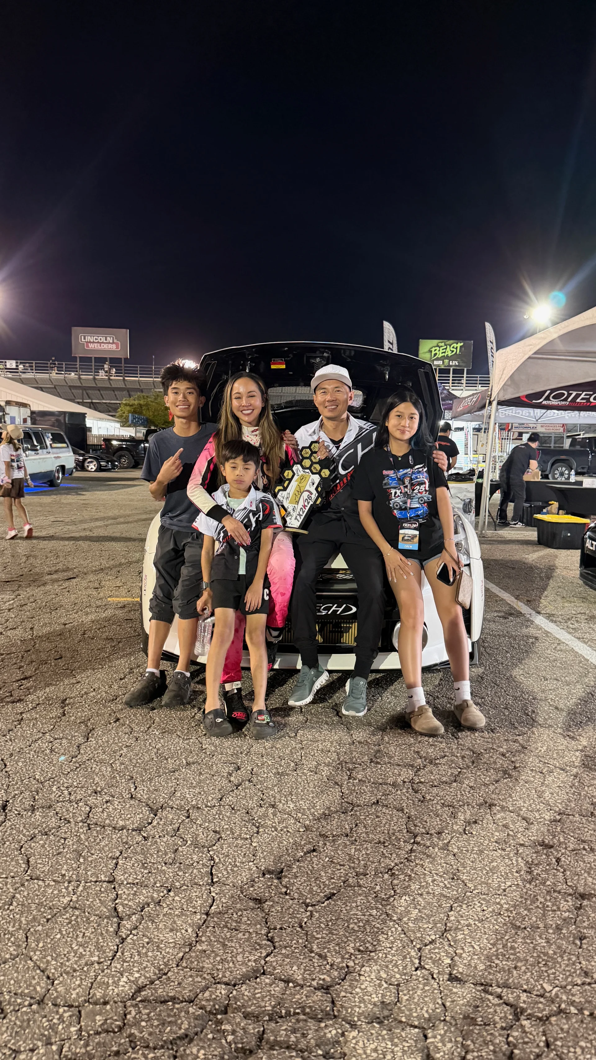 A family poses with trophies in the trunk of a white car at a night drag racing event.