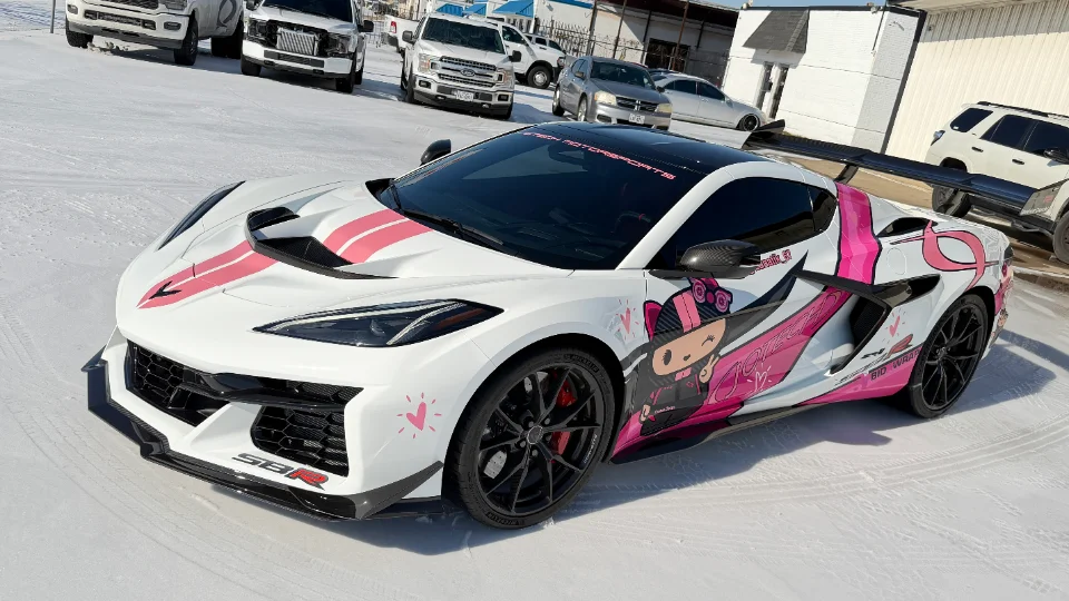 A white Corvette C8 with pink racing stripes and a custom anime-style wrap, featuring a large rear wing, parked on a snowy surface.