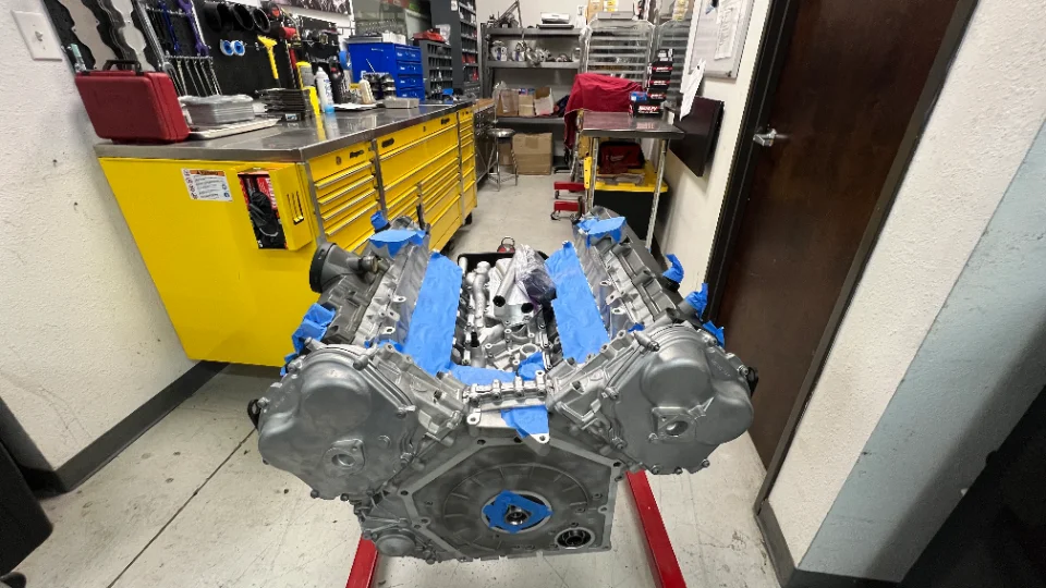 A V8 engine block on a stand in a workshop, with blue painter's tape protecting components during assembly.
