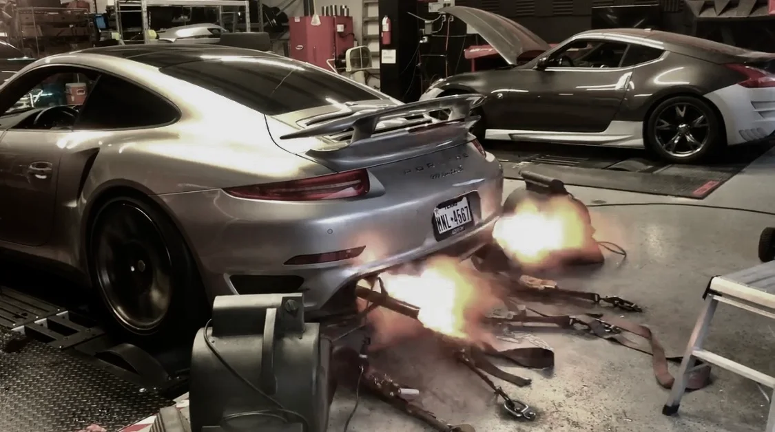 A silver Porsche 911 on a dyno with flames shooting from its exhaust during a performance tuning session, with another car in the background.