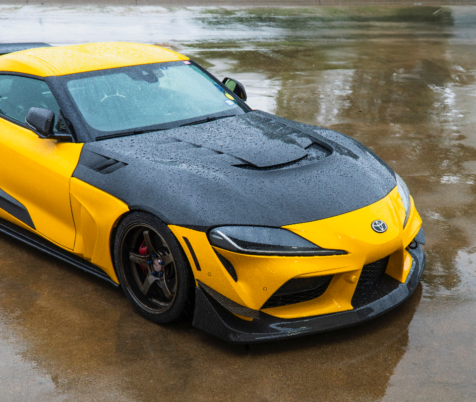 A modified yellow and black Toyota Supra with a carbon fiber hood and widebody kit parked on wet pavement.