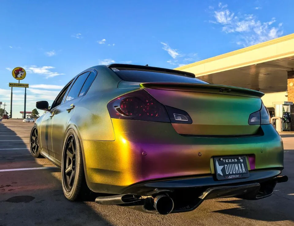 Rear view of a chameleon-wrapped Infiniti G37 sedan with aftermarket wheels, rear spoiler, and diffuser parked outdoors.
