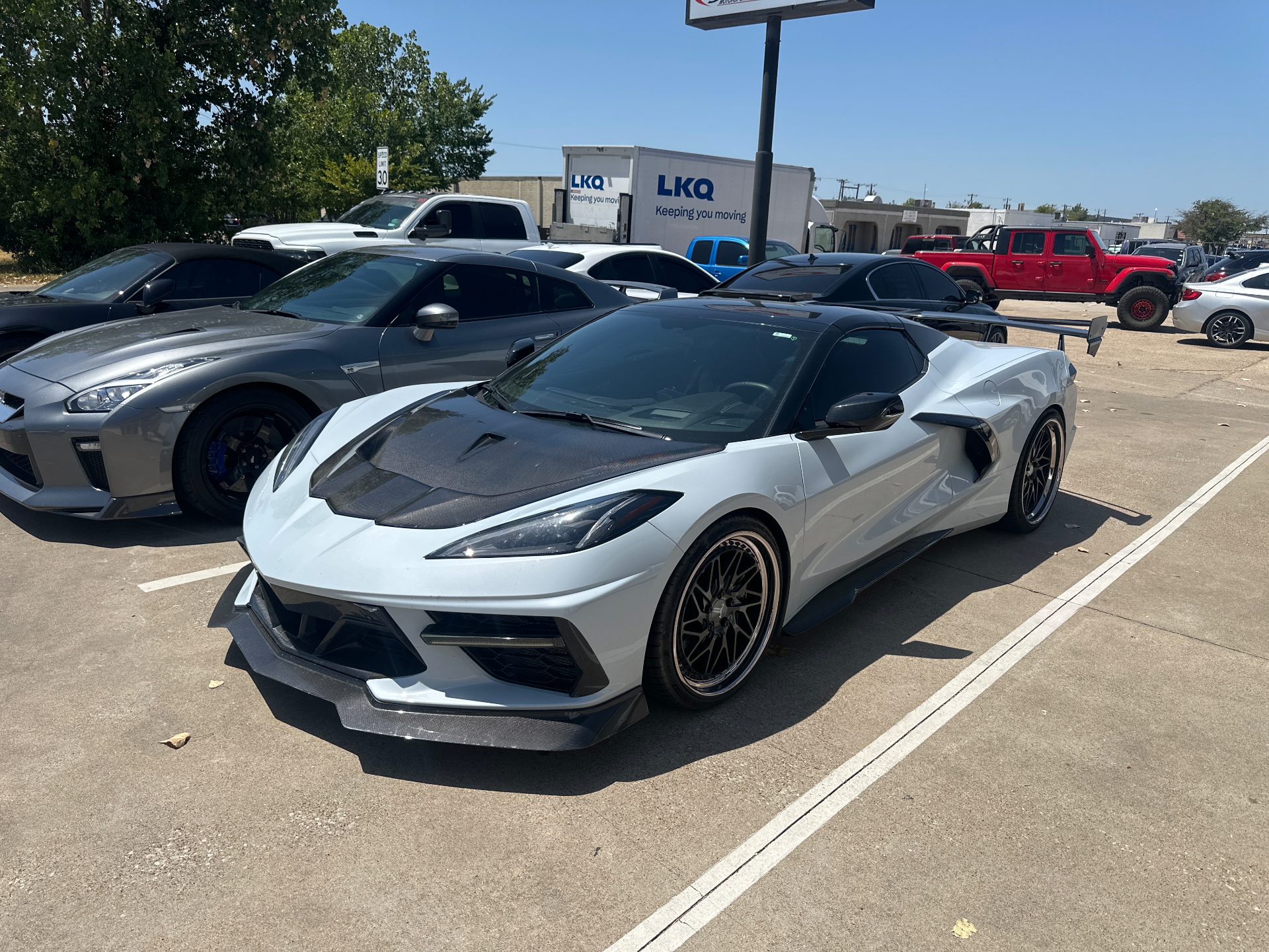 A modified white Chevrolet Corvette C8 with a carbon fiber hood and large rear wing parked next to a grey Nissan GT-R in a parking lot.