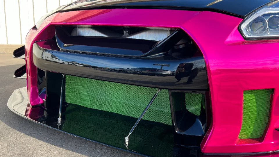 Close-up of a pink Nissan GT-R's front bumper, featuring a large splitter, canards, and a visible green intercooler.