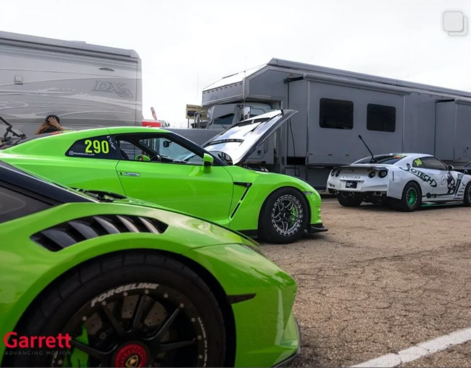 Three high-performance race cars, including a green Lamborghini and two Nissan GT-Rs, parked in a track paddock with trailers.