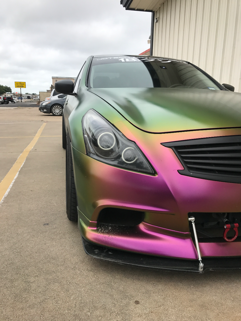 Front quarter view of a custom Infiniti G37 sedan with a vibrant chameleon wrap, front splitter, and red tow hook.