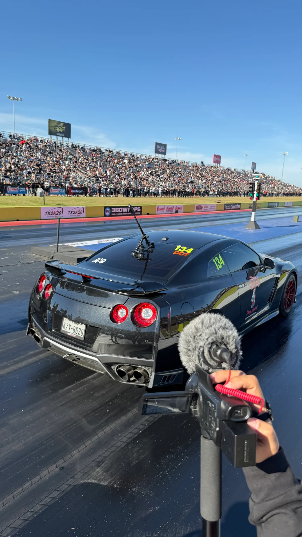 A black Nissan GT-R at the starting line of a drag strip with spectators in the stands, being filmed by a camcorder.