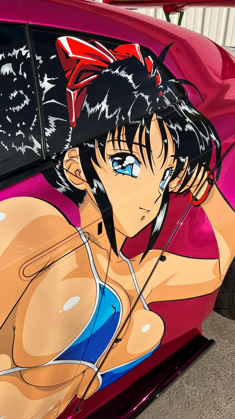 A close-up of a pink car with a large anime girl graphic wrap on the side, featuring a character with blue eyes and a red bow.