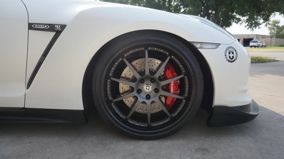 Close-up of a white Nissan GT-R's front wheel, showcasing HRE wheels and red Brembo brake calipers.