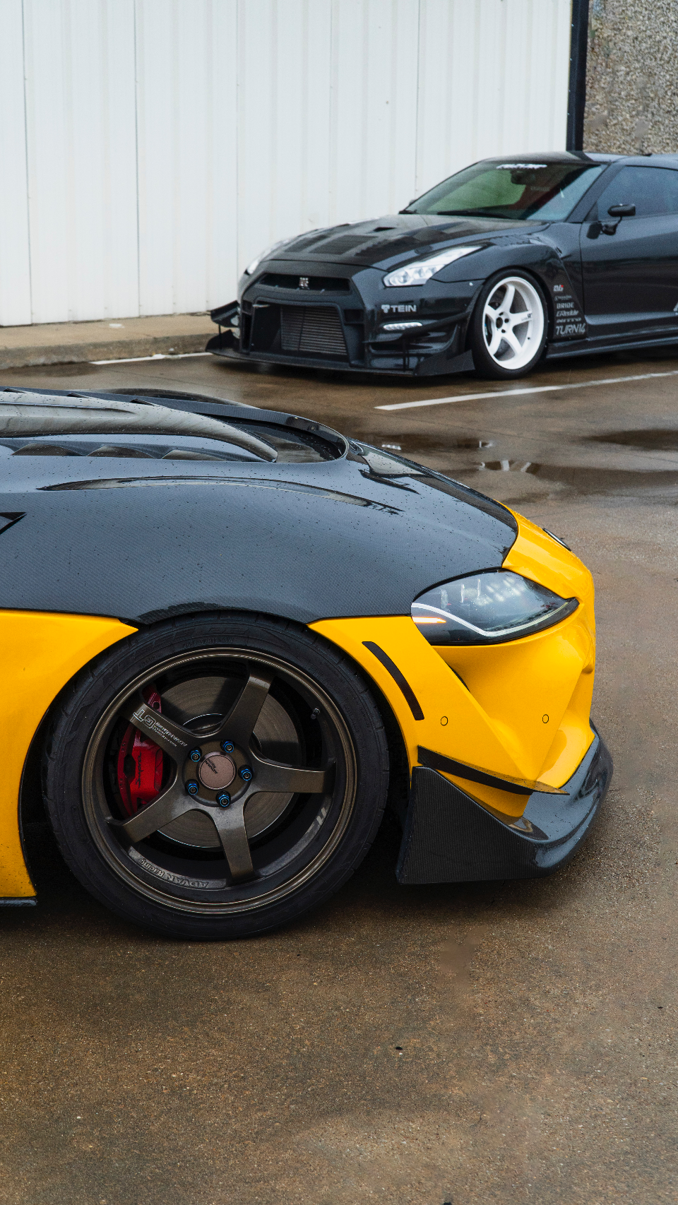 Two highly modified sports cars, a yellow and black car with carbon fiber hood and Advan wheels in the foreground, and a black Nissan GT-R with white wheels in 