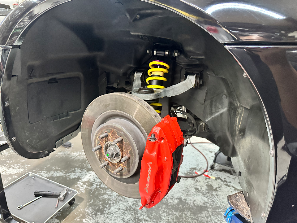 Close-up of a car's front wheel well in a shop, showing a red Viper brake caliper, large rotor, and yellow coilover suspension.