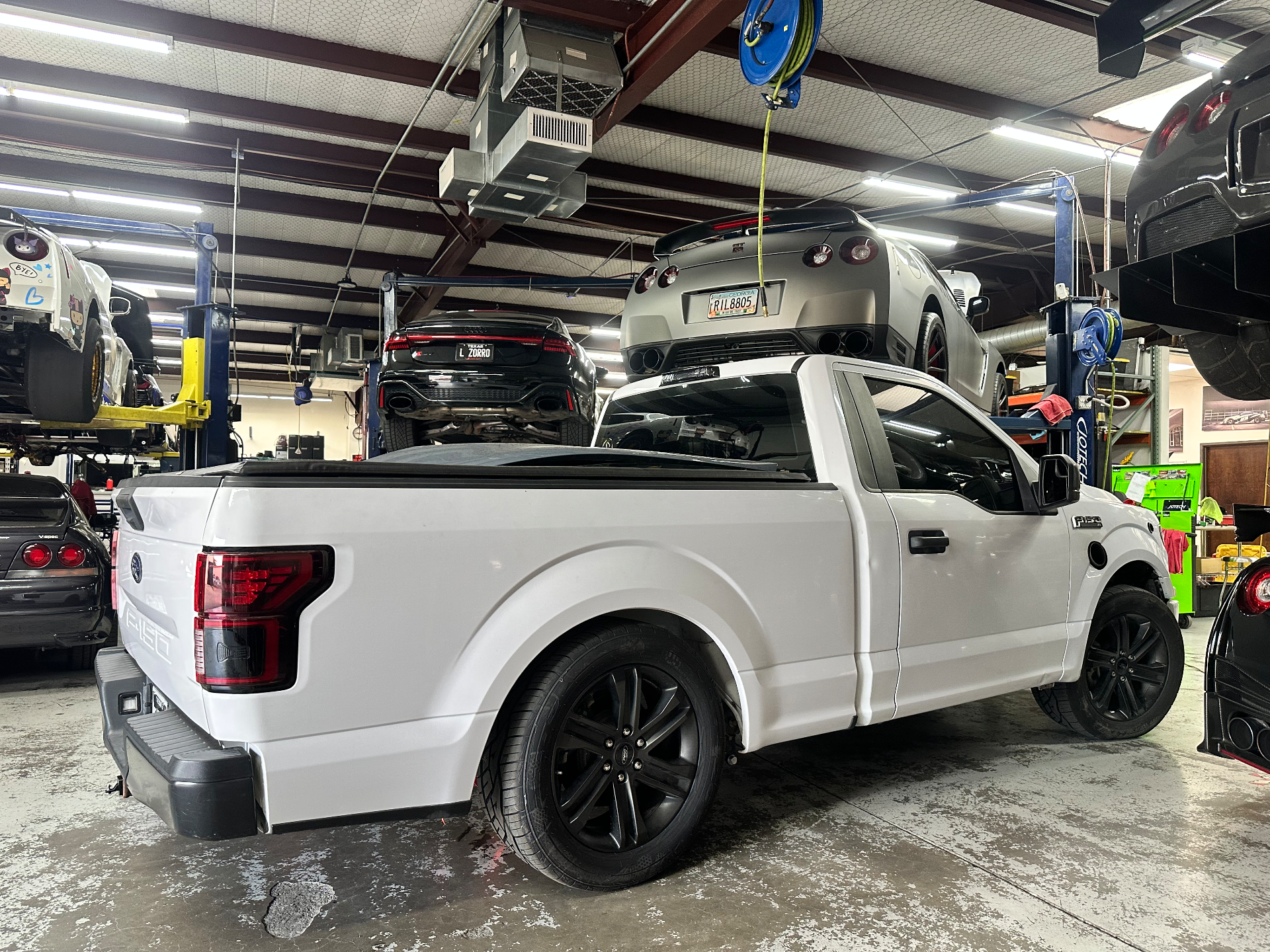 A white Ford F-150 truck on the floor of a performance tuning shop with multiple Nissan GT-Rs and an Audi RS6 on lifts.