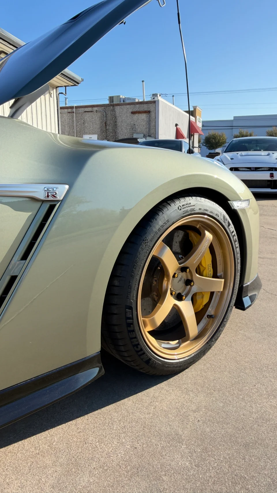 Close-up of a gold Nissan GT-R R35 wheel with Michelin tires and yellow brake calipers, parked outdoors with its hood open.