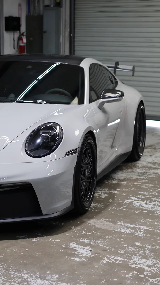 A white Porsche 911 GT3 with custom black multi-spoke wheels and a large rear wing parked in a garage.