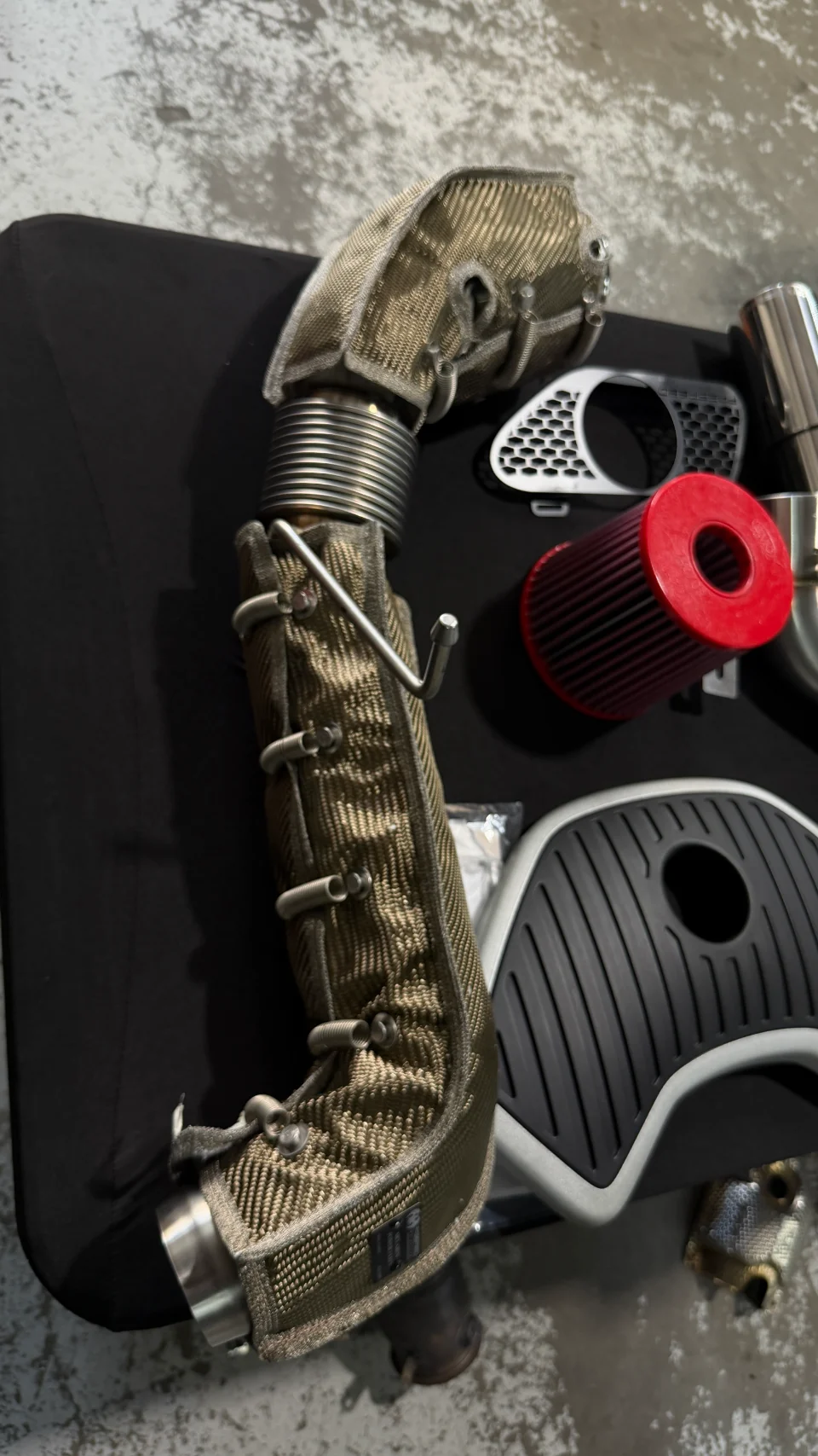 Aftermarket performance parts including a heat-wrapped downpipe, red air filter, and various engine covers laid out.
