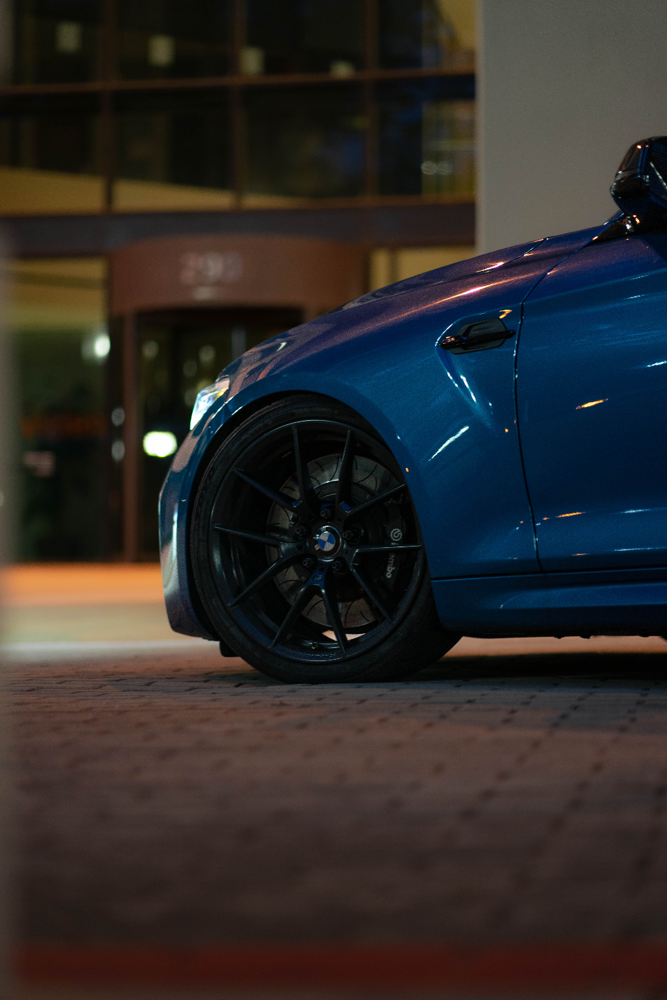 Close-up of a blue BMW M2's front wheel, featuring black multi-spoke wheels and Brembo brakes at night.